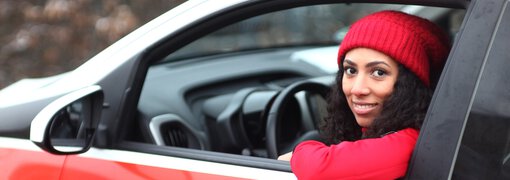Dunkelhaarige Frau mit langen locken und roter Mütze und Pulli schaut lächelnd aus der Fahrertür eines Caritas Pflege Auto heraus. | © heydenaber
